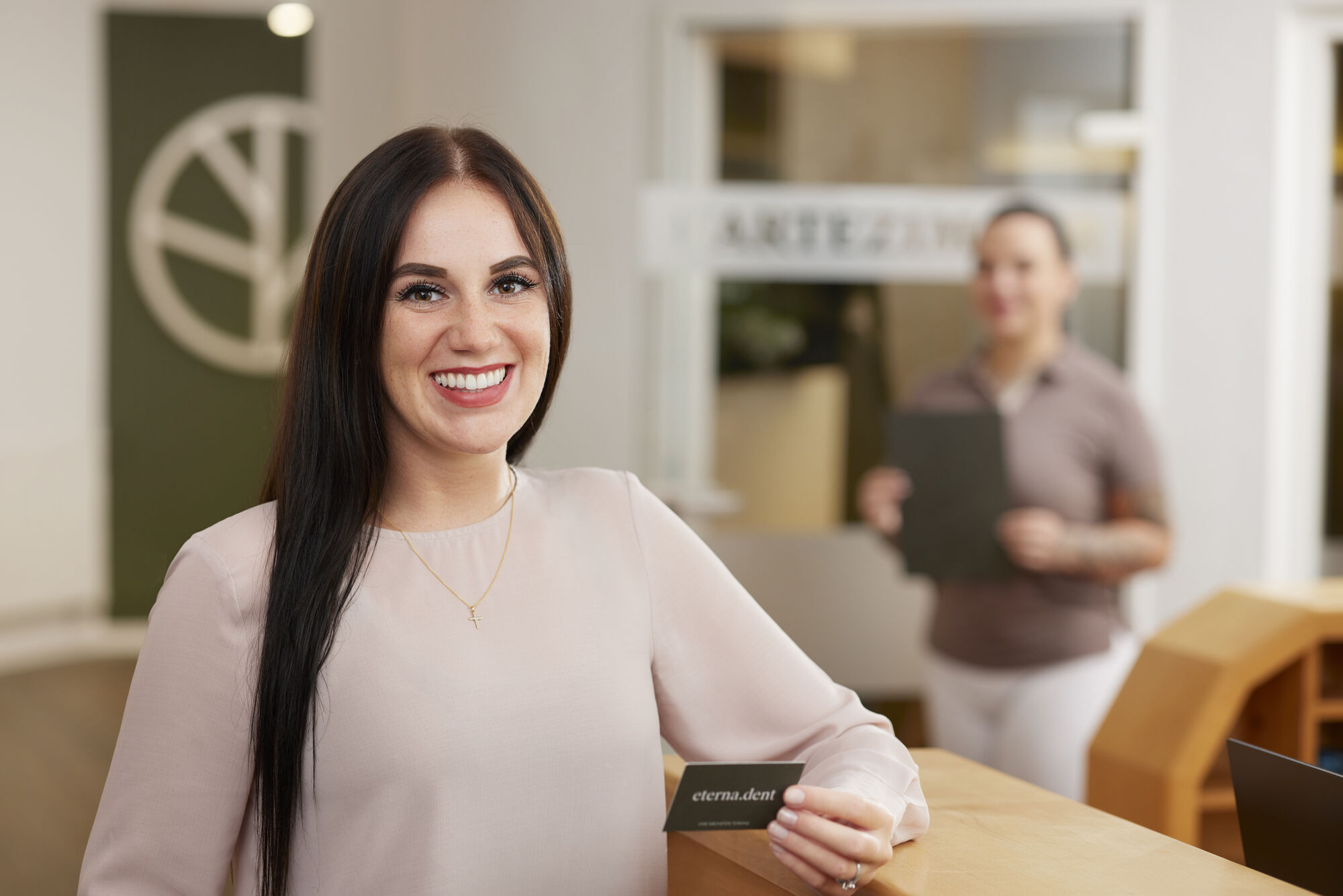 Eine Patientin der Praxis steht am Empfangstresen, hält eine Visitenkarte in der Hand und lächelt freundlich in die Kamera