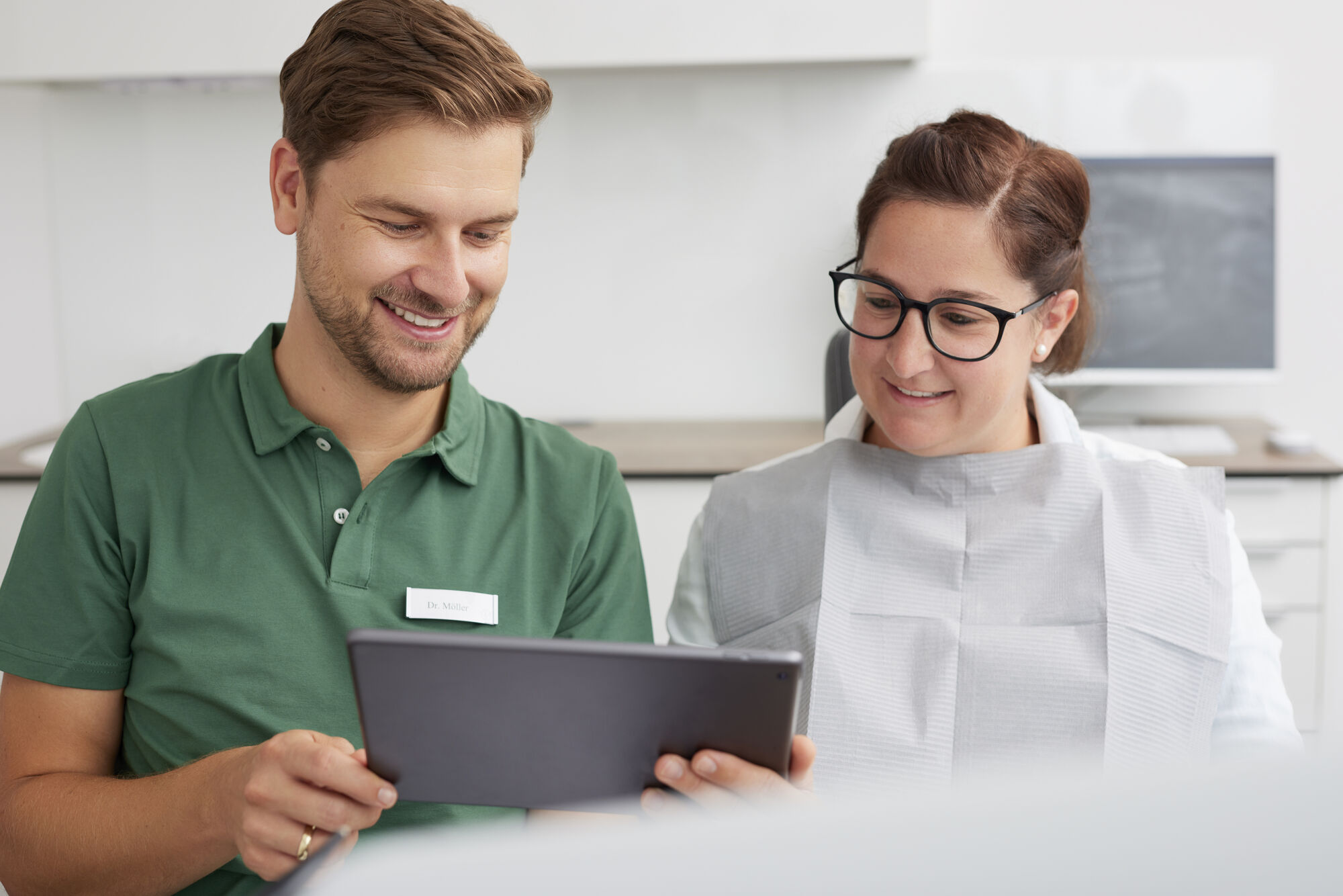 Dr. Möller zeigt einer Patientin Unterlagen zur Behandlung, beide lachen und sitzen nebeneinander im hellen und modernen Behandlungsraum