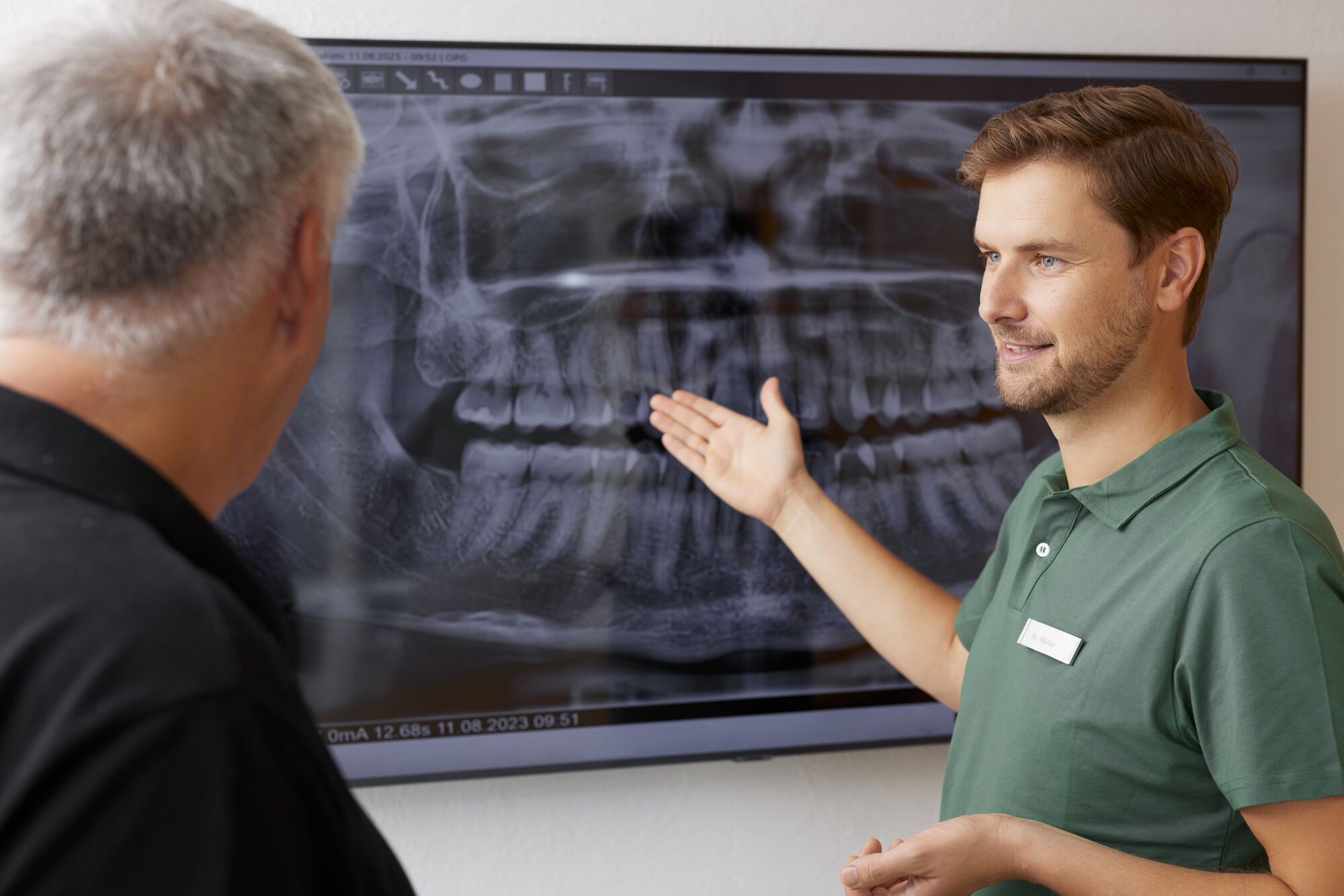 r_06A1140 Dr. Möller zeigt Patient ein großes Röntgenbild auf Monitor und erklärt Befunde, beide stehen im hellen Behandlungsraum der Zahnarztpraxis