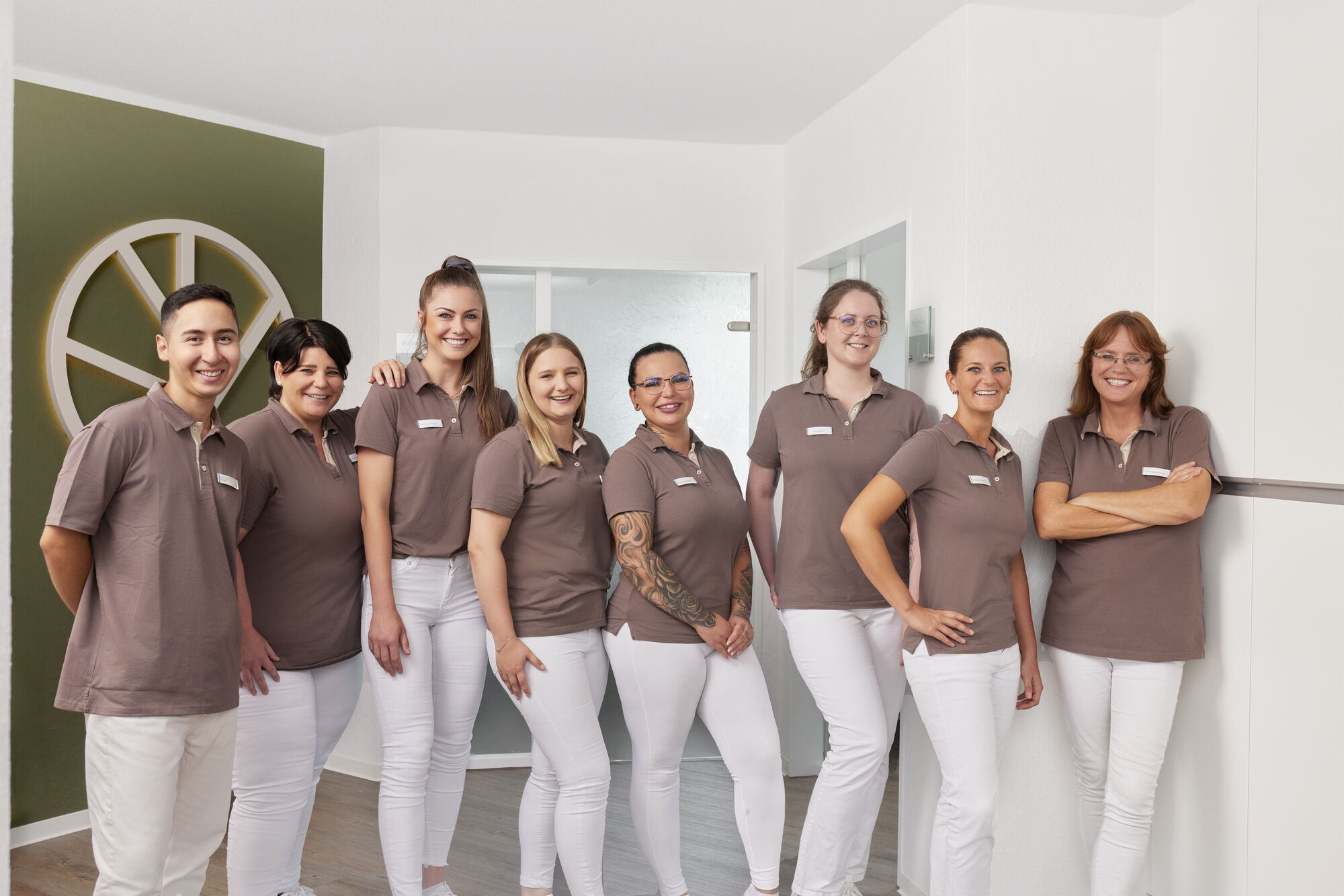 Teamfoto der Zahnarztpraxis eterna.dent: Mitarbeiterinnen in brauner Uniform und weißen Hosen stehen nebeneinander vor Praxislogo